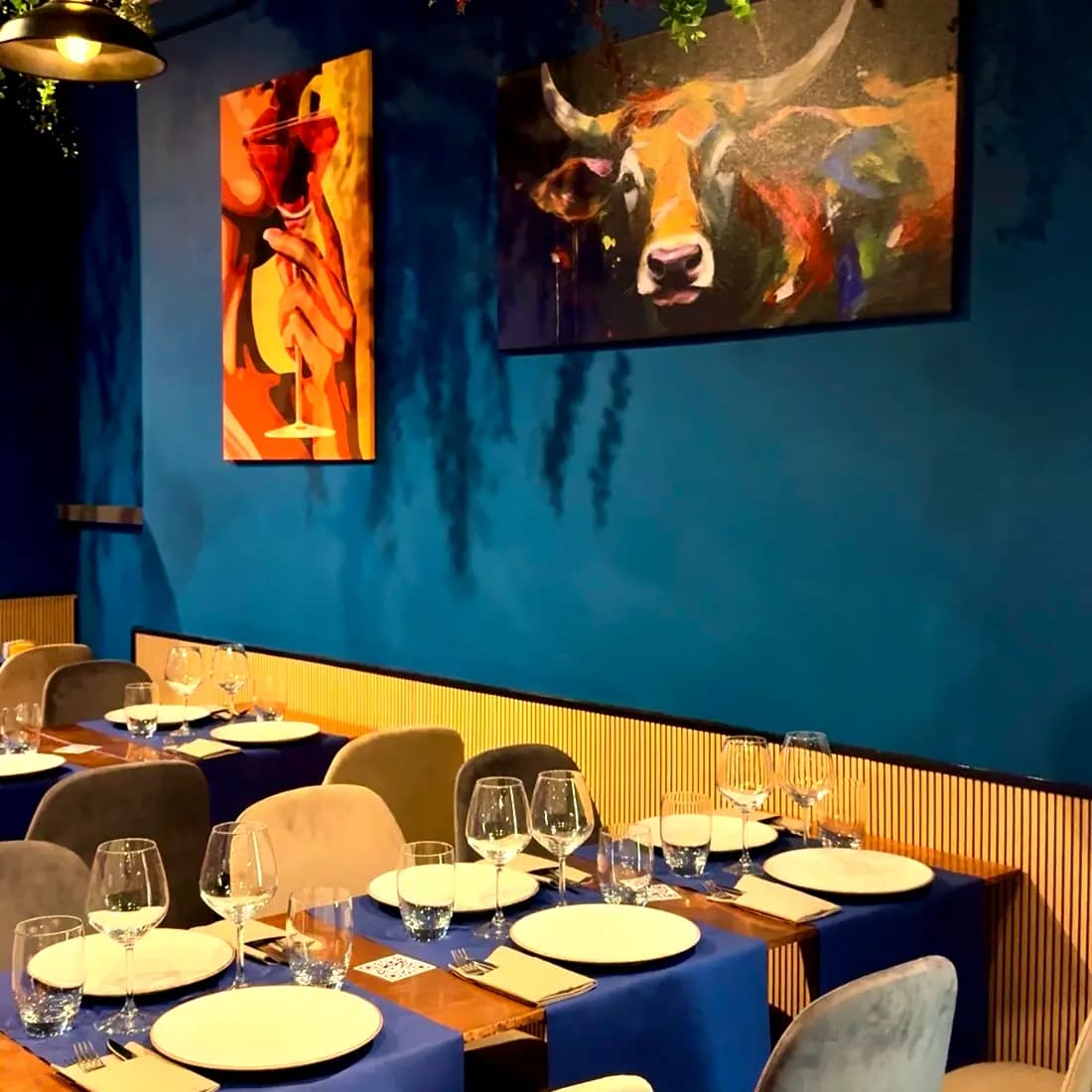 La sala di VinCarne by Dixie: tavoli apparecchiati con runner blu sotto i quadri del toro e dei calici, lungo la boiserie in legno